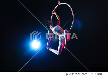 Young woman performing acrobatic element on aerial ring against dark background 95318013