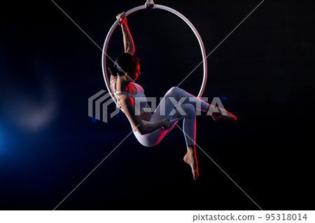 Young woman performing acrobatic element on aerial ring against dark background Young woman performing acrobatic element on aerial ring against dark background 95318014