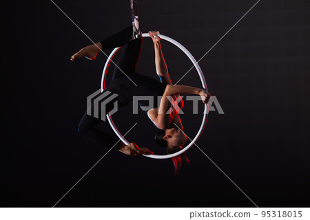 Young woman performing acrobatic element on aerial ring against dark background Young woman performing acrobatic element on aerial ring against dark background 95318015