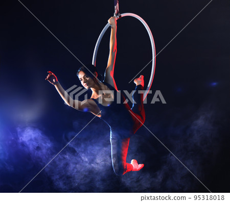 Young woman performing acrobatic element on aerial ring against dark background Young woman performing acrobatic element on aerial ring against dark background 95318018
