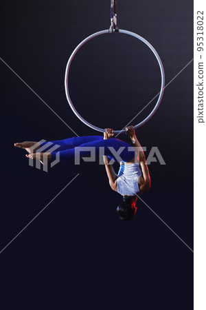 Young woman performing acrobatic element on aerial ring against dark background Young woman performing acrobatic element on aerial ring against dark background 95318022