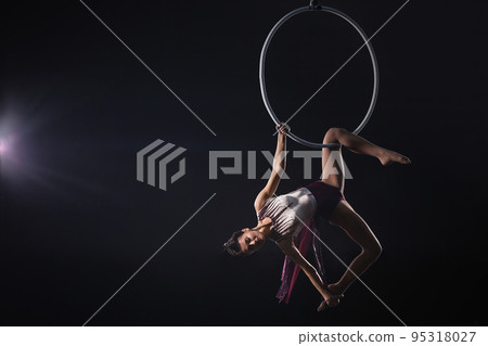 Young woman performing acrobatic element on aerial ring against dark background. Space for text 95318027