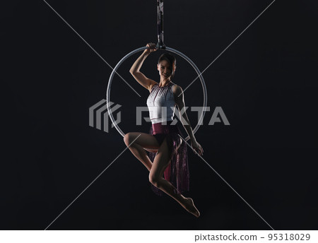 Young woman performing acrobatic element on aerial ring against dark background 95318029