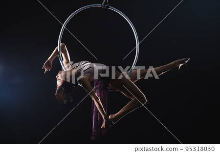 Young woman performing acrobatic element on aerial ring against dark background Young woman performing acrobatic element on aerial ring against dark background 95318030