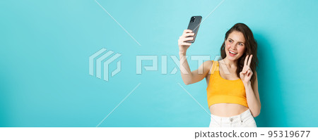 Technology and lifestyle concept. Stylish young woman taking selfie in her summer clothes, showing v-sign at smartphone camera, standing over blue background 95319677