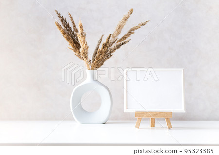 Empty picture frame mock up in minimalist interior with dried plant in vase on white table. Empty picture frame mock up in minimalist interior with dried plant in vase on white table. 95323385
