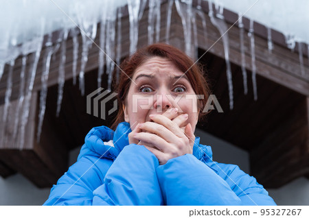 A frightened woman stands under a roof with large icicles. Danger in the winter season. A frightened woman stands under a roof with large icicles. Danger in the winter season. 95327267