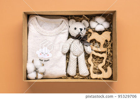 Gift basket with gender neutral baby garment and accessories. Care box of organic newborn cotton clothes, teddy, cotton in box on orange background Flat lay Top view. Gift basket with gender neutral baby garment and accessories. Care box of organic newborn cotton clothes, teddy, cotton in box on orange background Flat lay Top view. 95329706