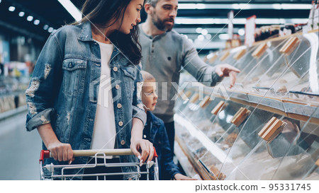 Mother, father and their little son are shopping for food in supermarket pointing at plastic boxes on shelves and talking. Large food store and its employees are visible in background. Mother, father and their little son are shopping for food in supermarket pointing at plastic boxes on shelves and talking. Large food store and its employees are visible in background. 95331745