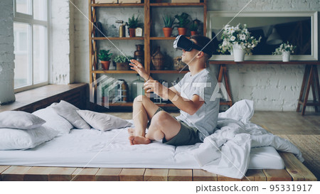 Excited young man in casual clothes is using augmented reality glasses moving hands while sitting on double bed in light apartment. Modern technology and happy people concept. 95331917