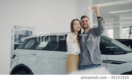 Attractive young couple is taking selfie with car keys in modern motor showroom after buying new automobile. They are looking at camera, posing, kissing and smiling. 95331920