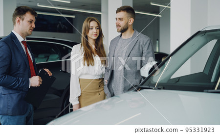 Friendly car salesman in suit is talking to confident young man telling him about new vehicle model while pretty woman is standing near her husband holding his arm and smiling. 95331923