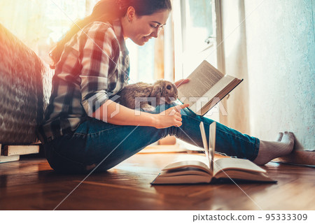 A happy young woman is sitting on the floor, holding a rabbit on her lap and showing it a book. Side view. The concept of reading and education A happy young woman is sitting on the floor, holding a rabbit on her lap and showing it a book. Side view. The concept of reading and education 95333309