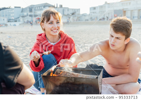 Group of friends sitting around the fire, grilling sausages and having great time on the beach picnic. Active Teenagers outdoor activity. Sunset time. Selective focus. Group of friends sitting around the fire, grilling sausages and having great time on the beach picnic. Active Teenagers outdoor activity. Sunset time. Selective focus. 95333713