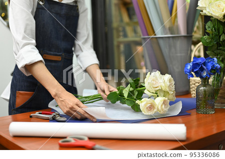 Female florist in apron is arranging, wrapping bouquet of white roses on a wooden table in craft paper Female florist in apron is arranging, wrapping bouquet of white roses on a wooden table in craft paper 95336086