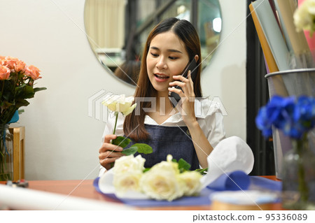 Pretty young woman talking on mobile phone, taking orders from clients while sitting in her floral shop Pretty young woman talking on mobile phone, taking orders from clients while sitting in her floral shop 95336089