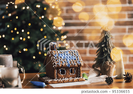 Little homemade gingerbread house standing on table with decorations, candles in living room decorated with defocused New Year lights, Christmas fir tree. Wonderful happy holiday mood. Copy space 95336655