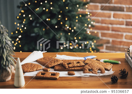 Homemade gingerbread house building blocks with glaze laying on table with decorations, lanterns in living room decorated with defocused New Year lights, Christmas fir tree. Holiday mood for kids Homemade gingerbread house building blocks with glaze laying on table with decorations, lanterns in living room decorated with defocused New Year lights, Christmas fir tree. Holiday mood for kids 95336657
