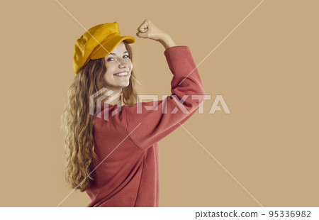 Cheerful pretty young woman in autumn beret hat flexing her strong arm and smiling 95336982