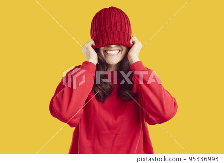 Joyful young woman covers her eyes with warm red winter hat that she puts on her head Joyful young woman covers her eyes with warm red winter hat that she puts on her head 95336993