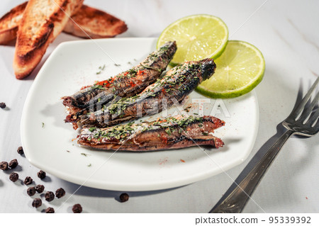 Appetizer of sardines in oil with parsley and paprika on a plate with some toast and lemon slices. 95339392