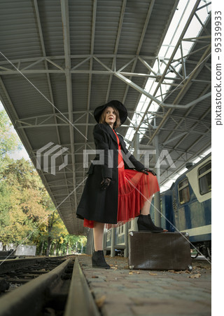 Portrait of young woman in red dress, black coat and hat on platform near train with leather suitcase. Vertical frame. Portrait of young woman in red dress, black coat and hat on platform near train with leather suitcase. Vertical frame. 95339933