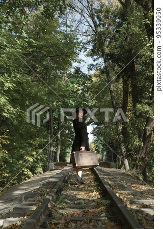 Woman in black clothes leaves with a suitcase along the rails through the autumn forest. Back view 95339950