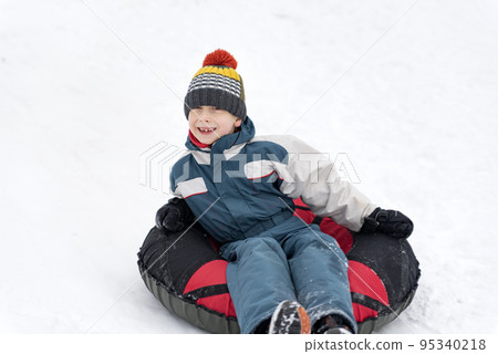 Happy boy in winter jumpsuit and hat with bubo rides on tubing in snow. Merry snowy winter fun Happy boy in winter jumpsuit and hat with bubo rides on tubing in snow. Merry snowy winter fun 95340218