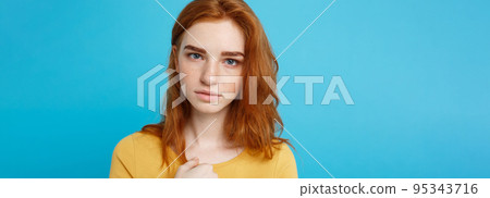 Headshot Portrait of tender redhead teenage girl with serious expression looking at camera. Caucasian woman model with ginger hair posing indoors.Pastel blue background. Copy Space. 95343716
