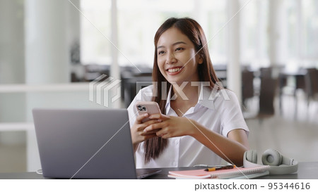 Smiling Asian beautiful woman using mobile smartphone while working with laptop computer. 95344616