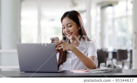 Smiling Asian beautiful woman using mobile smartphone while working with laptop computer. Smiling Asian beautiful woman using mobile smartphone while working with laptop computer. 95344617