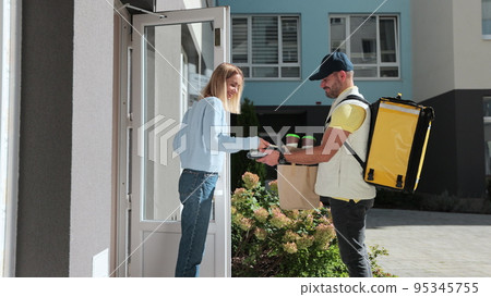 Woman open door to a delivery man on a bike wearing yellow thermal backpack with POS wireless terminal for smartphone paying and recieves coffee and food in paper bag. Food delivery concept. Woman open door to a delivery man on a bike wearing yellow thermal backpack with POS wireless terminal for smartphone paying and recieves coffee and food in paper bag. Food delivery concept. 95345755