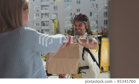 Woman open door to a food delivery man on a bike wearing yellow thermal backpack and recieves order or package with food. Food delivery concept. 95345760