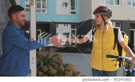 Close up of handsome man open door to a pizza delivery woman wearing yellow thermal backpack on a bike and recieves cardboard boxes of pizza. Food delivery concept. 95345801