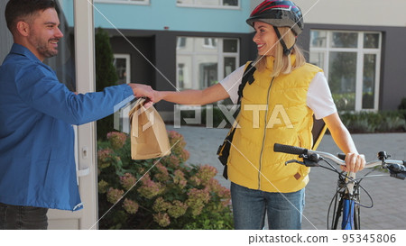 Close up man customer open door to a food delivery woman wearing yellow thermal backpack on a bike and recieves order or package with food. Food delivery concept. Close up man customer open door to a food delivery woman wearing yellow thermal backpack on a bike and recieves order or package with food. Food delivery concept. 95345806