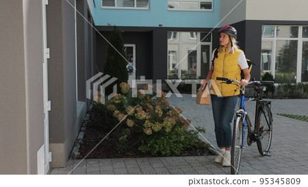 Man customer open door to a food delivery woman wearing yellow thermal backpack on a bike and recieves order or package with food. Food delivery concept. 95345809