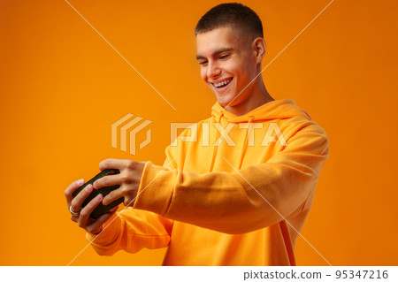 Portrait of excited young man playing online game on mobile phone on yellow background. 95347216