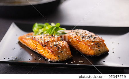 Two salmon fillets baked until crispy, sprinkled with sesame on a black plate. 95347759
