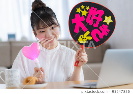 Oshikatsu A young woman who cheers for the Oshi by waving a fan while watching a live video 95347826