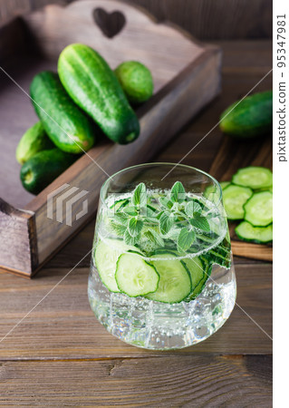 Detox drink. Water with pieces of cucumber, ice and mint leaves in a transparent glass on a wooden table. Vertical view Detox drink. Water with pieces of cucumber, ice and mint leaves in a transparent glass on a wooden table. Vertical view 95347981