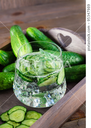 Detox drink. Water with pieces of cucumber, ice and mint leaves in a transparent glass on a wooden tray on the table Detox drink. Water with pieces of cucumber, ice and mint leaves in a transparent glass on a wooden tray on the table 95347989