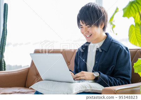 A man in his 20s working on a computer with a smile-living room sofa and foliage plants A man in his 20s working on a computer with a smile-living room sofa and foliage plants 95348823