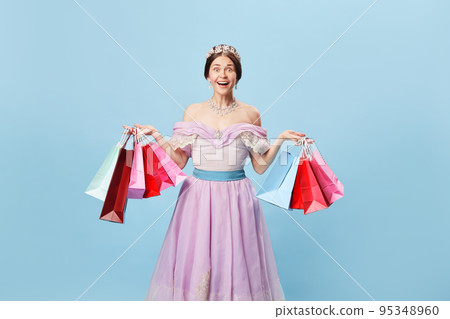 Excited princess. Young woman in image of young queen holding bright shopping packages looking at camera with delight isolated on blue background. Excited princess. Young woman in image of young queen holding bright shopping packages looking at camera with delight isolated on blue background. 95348960