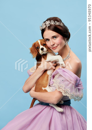 Beloved pet. Portrait of young charming woman in lilac color vintage dress as medieval princess holding small King Charles spaniel dog on blue background. 95348970