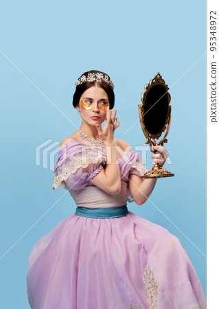Face skin care. Beautiful charming woman in lilac color vintage dress as a royal person, princess looking in mirror at herself on blue background. Face skin care. Beautiful charming woman in lilac color vintage dress as a royal person, princess looking in mirror at herself on blue background. 95348972