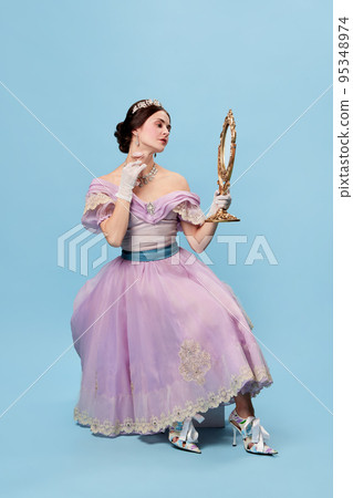 Young and pretty. Beautiful charming woman in lilac color vintage dress as a royal person, princess looking in mirror at herself on blue background. 95348974