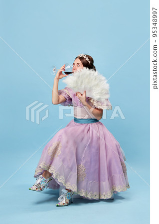 Tasting wine. Cute young girl in lilac color medieval dress as young queen or princess on blue background. Eras comparison, beauty, art, emotions and vintage fashion style Tasting wine. Cute young girl in lilac color medieval dress as young queen or princess on blue background. Eras comparison, beauty, art, emotions and vintage fashion style 95348997