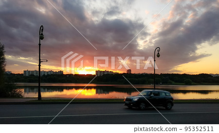 Landscape with sunset and dark clouds reflecting in the water, city buildings, road, rushing car  95352511