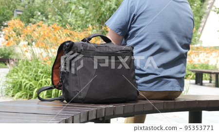 Bag is on park bench next to its owner. Man is resting with photo bag next to him. 95353771
