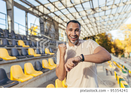 Happy young sportsman looking at camera and smiling happy with good fitness result, man checking time on fitness bracelet smart watch, running in sports stadium in daytime. 95354241